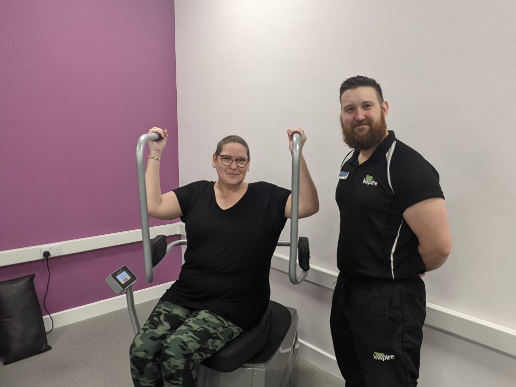 Dawn sits using some of the Wellness Hub equipment with staff member Kieran standing next to her.