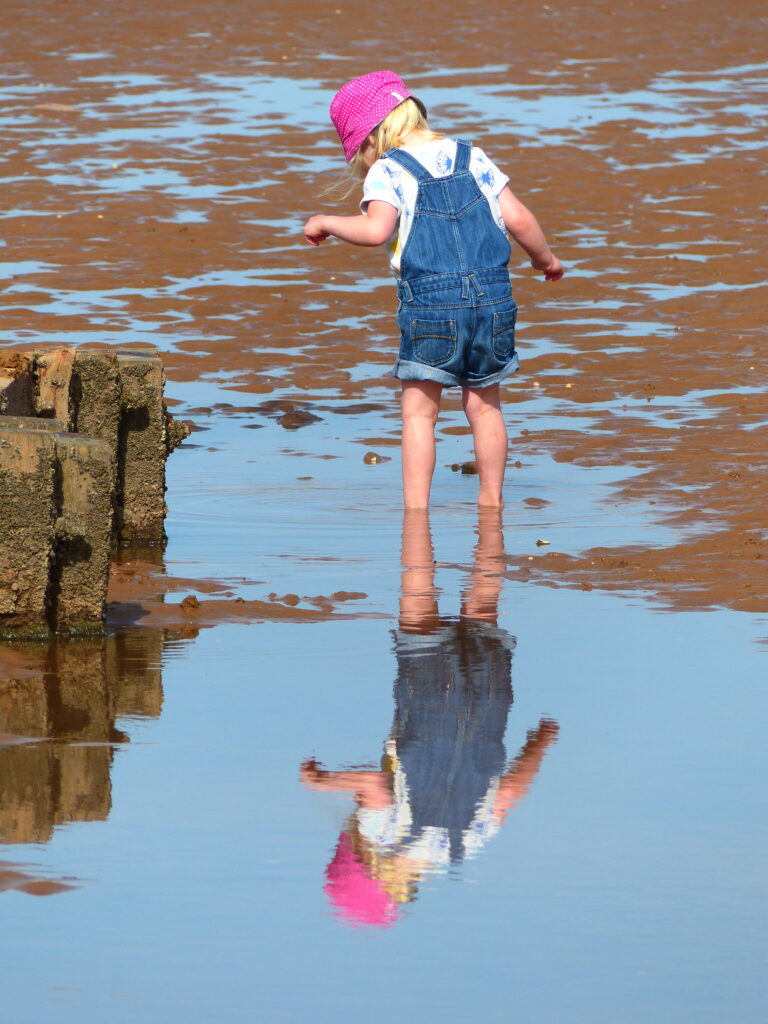 Girl in pink hat and dungarees looks into her reflection on the water on the beach