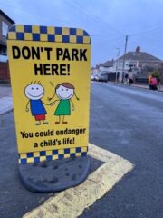 A sign asking parents not to park near schools with two small cartoon children on it holding hands