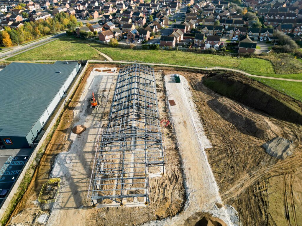 A photo of a steel frame of a primary school, standing next to a supermarket, with houses in the backgrounds.