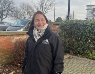 A picture of Liz, the rough sleeper coordinator for NELC. She has short brown hair, is wearing a white check scarf wrapped tightly round her and a black jacket with the NELC logo on the left.