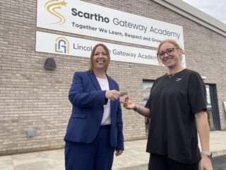 A photo of two people handing over keys in front of a school sign